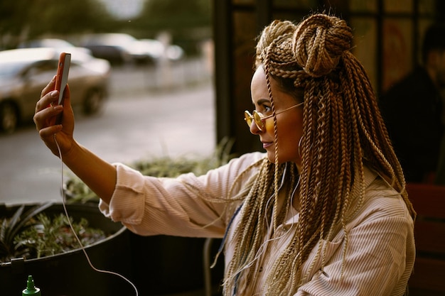 La giovane bella donna multirazziale con l'acconciatura afro bionda intreccia zizi sul caffè di strada con le cuffie. occhiali da sole gialli, trucco luminoso, stile hippie. videochiamata, lavoro fuori casa