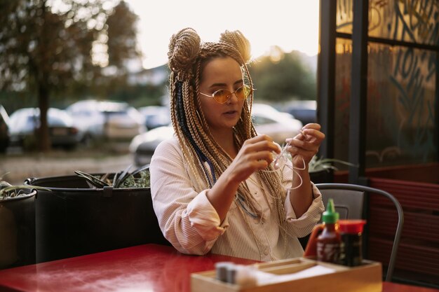 La giovane bella donna multirazziale con l'acconciatura afro bionda intreccia zizi sul caffè di strada con le cuffie. occhiali da sole gialli, trucco luminoso, stile hippie