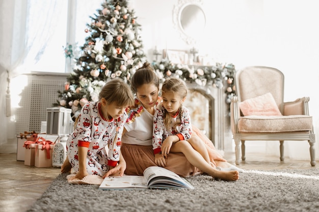 La giovane bella madre con due piccole figlie si siede sul tappeto e legge il libro vicino all'albero di Capodanno con i regali nella stanza luminosa e accogliente con poltrona e camino. .