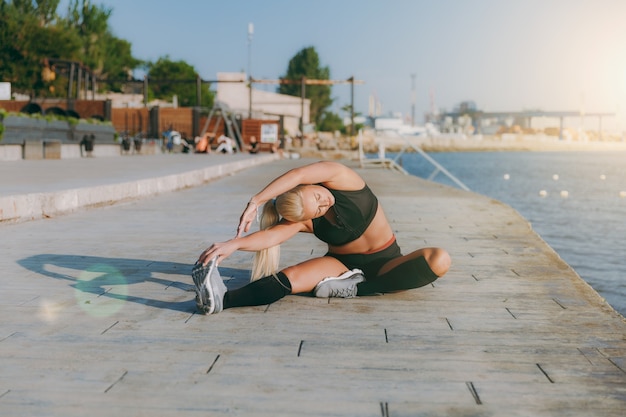 La giovane bella ragazza atletica con lunghi capelli biondi in abiti neri che si allena e fa esercizi all'alba sul mare