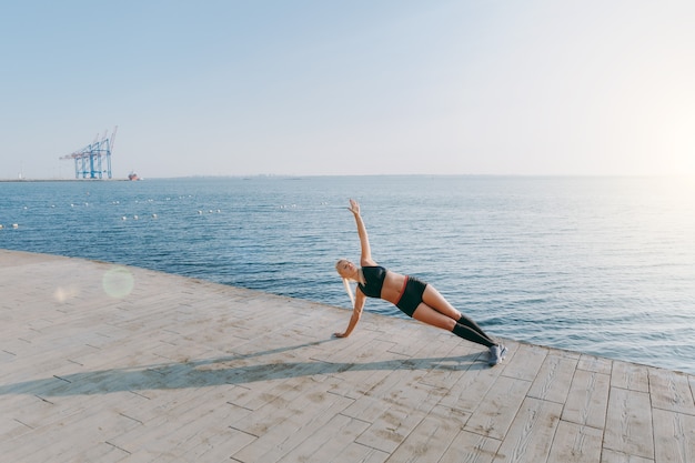 La giovane bella ragazza atletica con lunghi capelli biondi in abiti neri che si allena e fa esercizi, alzando il braccio all'alba sul mare