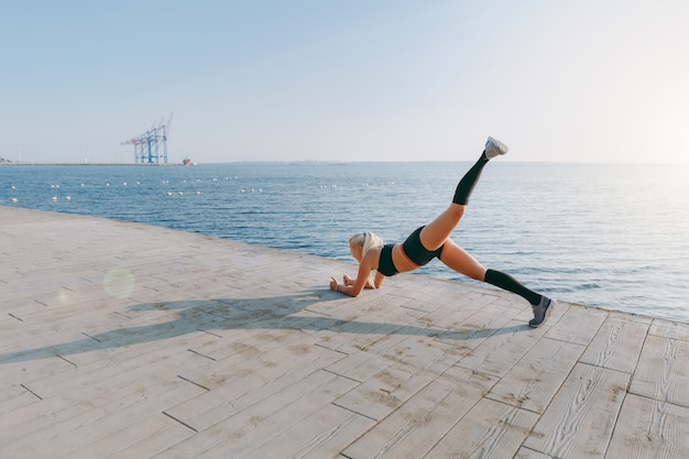 La giovane bella ragazza atletica con lunghi capelli biondi in abiti neri che si allena e fa esercizi, alzando la gamba all'alba sul mare