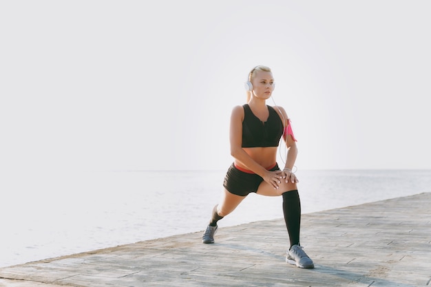 La giovane bella ragazza atletica con lunghi capelli biondi in cuffia e telefono cellulare in mano ascoltando musica e facendo stretching all'alba sul mare