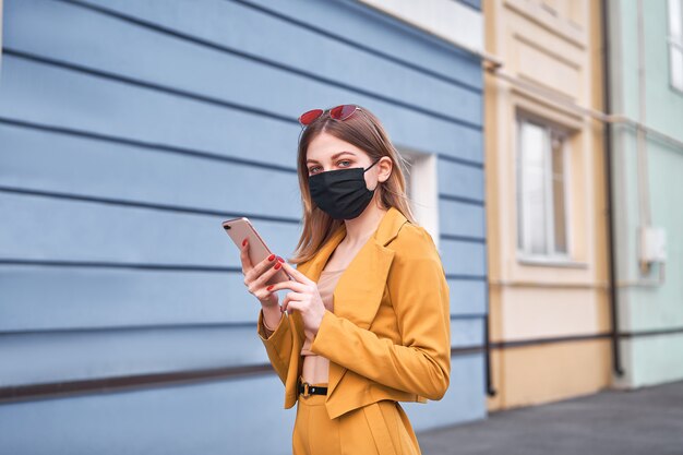 La giovane bella ragazza caucasica di 20 anni indossa una maschera nera contro l'epidemia di coronavirus covid-19 cammina per la strada e guarda lo schermo del telefono.