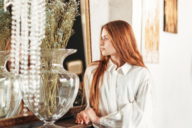 La giovane bella ragazza con capelli lunghi sta vicino allo specchio di mattina nella camera da letto