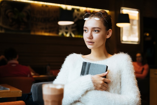 La giovane bella ragazza freelance con gli occhiali e un maglione bianco si siede in un caffè, tiene un taccuino per lavoro