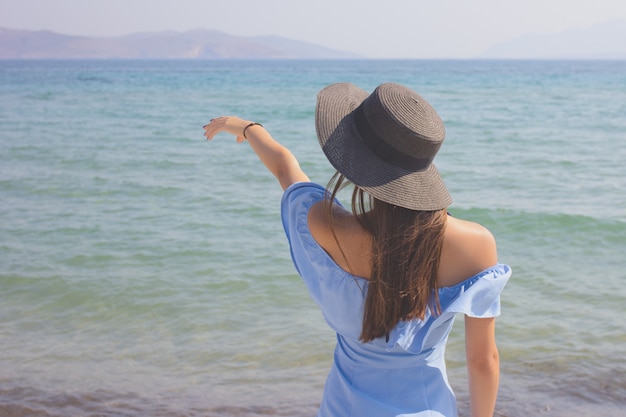 La giovane bella ragazza in un cappello guarda il mare, mostra una mano