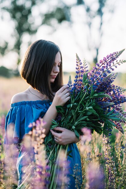 La giovane bella ragazza in un vestito sta in un campo di lupini. La ragazza tiene un grande mazzo di lupini viola in un campo di fioritura. Fiori di lupino in fiore. ecologico. Concetto di natura