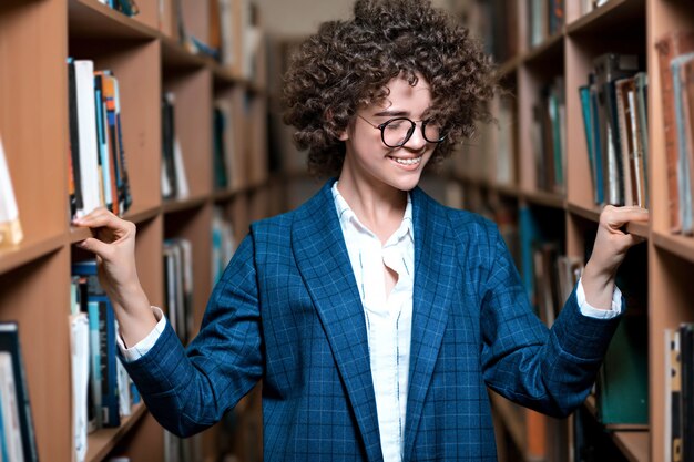 La giovane bella ragazza riccia in vetri e un vestito blu sta stando nella biblioteca