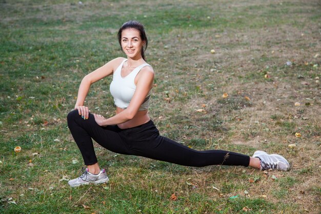 La giovane bella ragazza sexy felice sta allungando nel giardino del parco. sfondo di persone. con i capelli scuri e tuta sportiva in bianco e nero e felicità e sorriso. nella stagione estiva o autunnale.