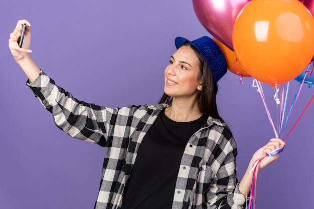 La giovane bella ragazza sorridente che indossa il cappello del partito che tiene i palloni prende un selfie