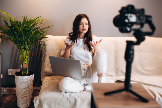 La giovane blogger femminile sorpresa è seduta sul divano con il laptop che registra il suo vlog di discorso al suo pubblico. Blogging indoor.