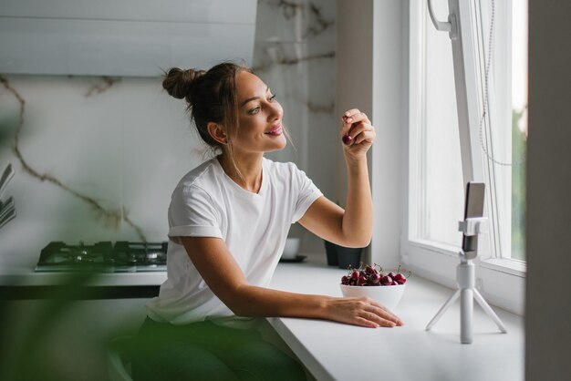 La giovane blogger nutrizionista conduce una consultazione o una lezione aperta tramite collegamento video che racconta i benefici delle bacche fresche sedute in cucina con un piatto di ciliegie