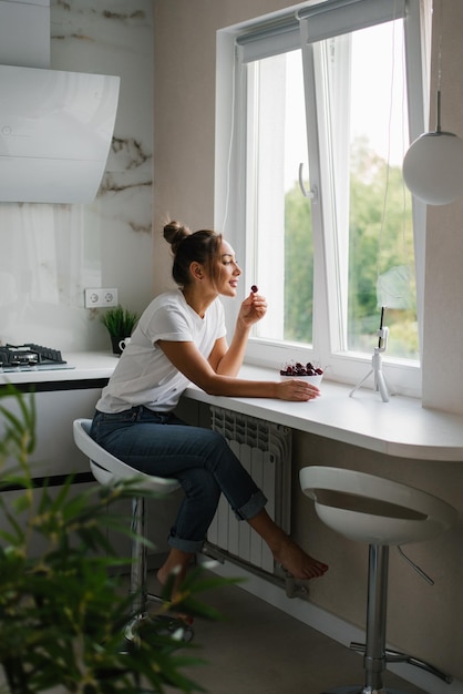 La giovane blogger nutrizionista conduce una consultazione o una lezione aperta tramite collegamento video che racconta i benefici delle bacche fresche sedute in cucina con un piatto di ciliegie