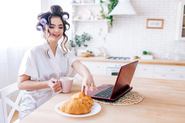 La giovane cameriera spensierata attraente fa colazione nella mattina in cucina. Raggiungere il cornetto. Laptop sul tavolo. Giovane donna che indossa i bigodini in capelli e vestaglia bianca.