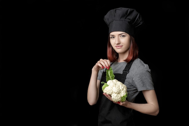 La giovane chef donna attraente in uniforme nera tiene il cavolfiore fresco su sfondo nero