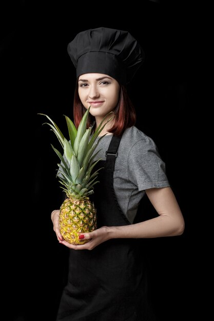 La giovane chef donna attraente in uniforme nera tiene l'ananas fresco su sfondo nero