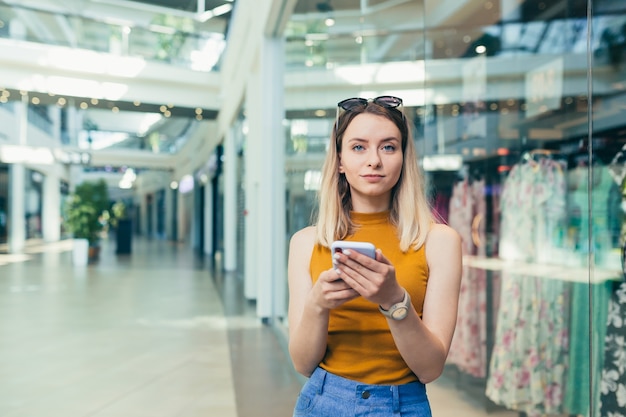 La giovane consumatrice nel centro commerciale naviga in chat e usa uno smartphone. femmina in piedi con un telefono cellulare in mano nel centro commerciale. interno. la ragazza felice dell'acquirente con i sacchetti regalo fa acquisti