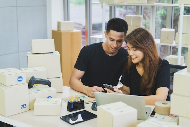 La giovane coppia asiatica felice di affari è lavoro insieme usando il computer portatile, lo smartphone e la compressa con una scatola del pacchetto che imballa al loro Ministero degli Interni startup, al venditore di affari online della PMI ed al concetto di consegna