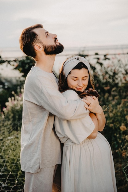 La giovane coppia autentica innamorata di un uomo e una donna vestiti con abiti di lino sono fotografati al tramonto in piedi tra i letti di fiori di campo su un effetto di rumore del fuoco selettivo di una fattoria di fiori