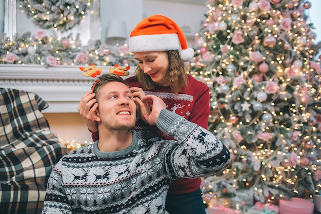 La giovane coppia è insieme. La donna sta dietro l'uomo e tiene le mani sulla sua testa. La guarda e le tocca la mano. Sono nella stanza decorata vicino al camino e all'albero di Natale.