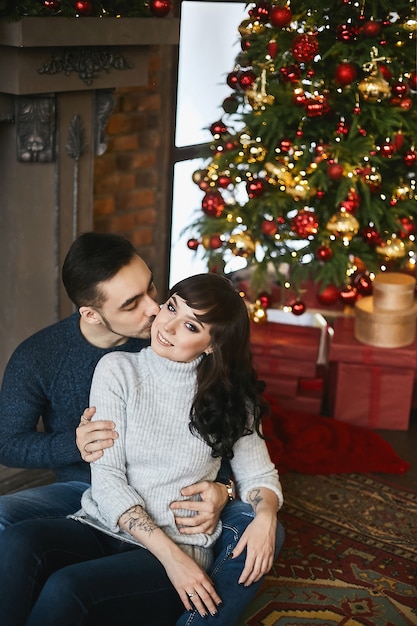 La giovane coppia felice, in caldi maglioni. Giovane uomo bello che bacia una bella ragazza vicino al camino e albero di Natale in background. Celebrazione del Natale e vacanze di Capodanno.