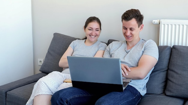 La giovane coppia, l'uomo e la donna stanno guardando un film comico sul portatile e stanno ridendo seduti sul divano. Sono nel loro appartamento moderno.