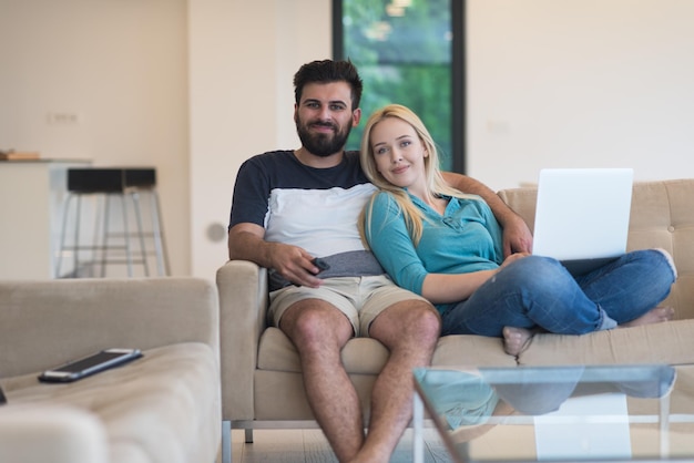 La giovane coppia si rilassa sul divano nel soggiorno di lusso, utilizzando un computer portatile e un telecomando