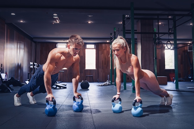 La giovane coppia sta lavorando in palestra. Donna attraente e bell'uomo muscoloso si stanno allenando nella moderna palestra. Fare plank con il kettlebell. Push-up sui pesi.