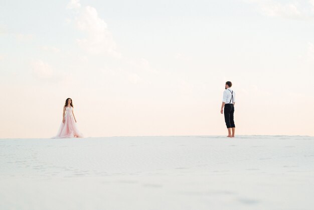 La giovane coppia un ragazzo in calzoni neri e una ragazza in un vestito rosa stanno camminando lungo la sabbia bianca del deserto