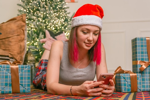 La giovane donna affascinante e felice giace sul pavimento e comunica al telefono sullo sfondo di un albero di Natale con regali