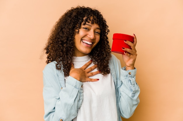 La giovane donna afroamericana afro che tiene un regalo di San Valentino ride ad alta voce tenendo la mano sul petto.