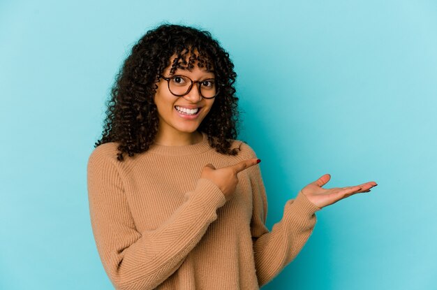 La giovane donna afroamericana afro isolata ha eccitato che tiene uno spazio della copia sul palmo.