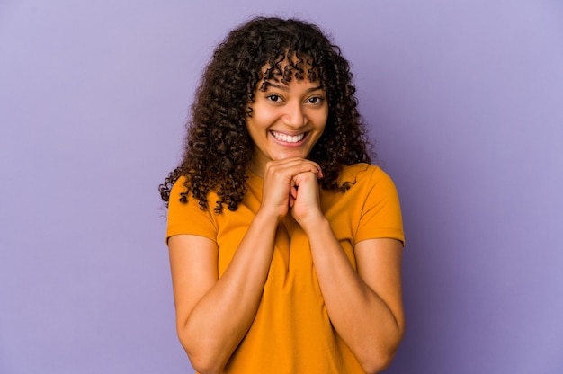 La giovane donna afroamericana afro isolata tiene le mani sotto il mento, sta guardando felicemente da parte.