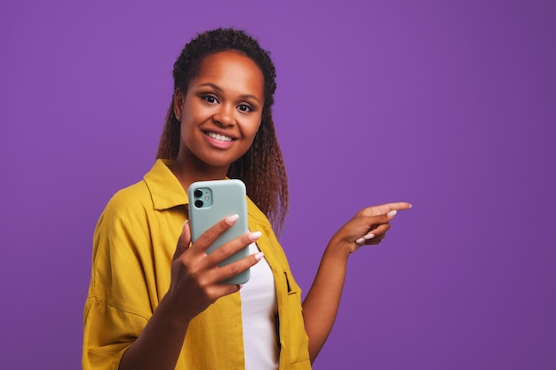 La giovane donna afroamericana casuale sorridente con il telefono punta il dito a lato