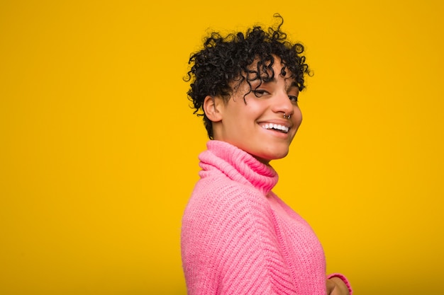 La giovane donna afroamericana che indossa un maglione rosa guarda da parte sorridente, allegro e piacevole.