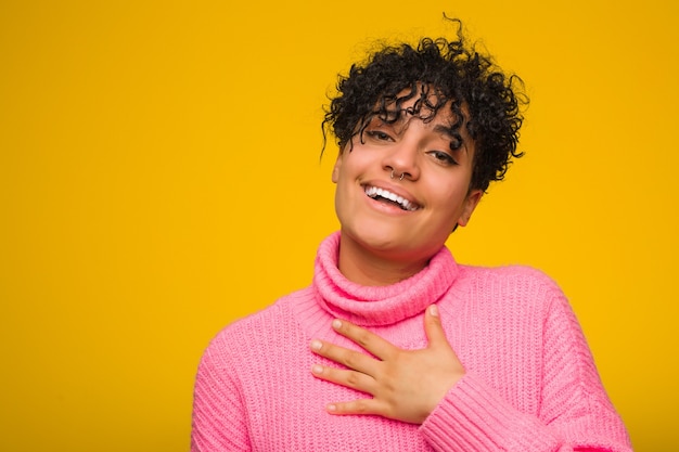 La giovane donna afroamericana che indossa un maglione rosa ride ad alta voce tenendo la mano sul petto.