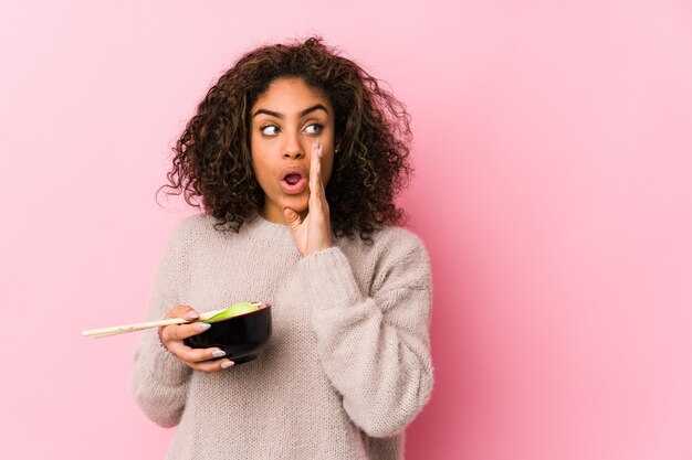 La giovane donna afroamericana che mangia le tagliatelle sta dicendo una notizia di frenata calda segreta e sta guardando da parte