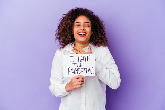 La giovane donna afroamericana che tiene io odio il cartello pandemico isolato sul muro viola ride ad alta voce tenendo la mano sul petto.