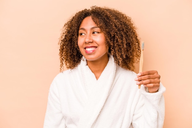 La giovane donna afroamericana che tiene lo spazzolino da denti isolato su sfondo beige sembra da parte sorridendo allegra e piacevole