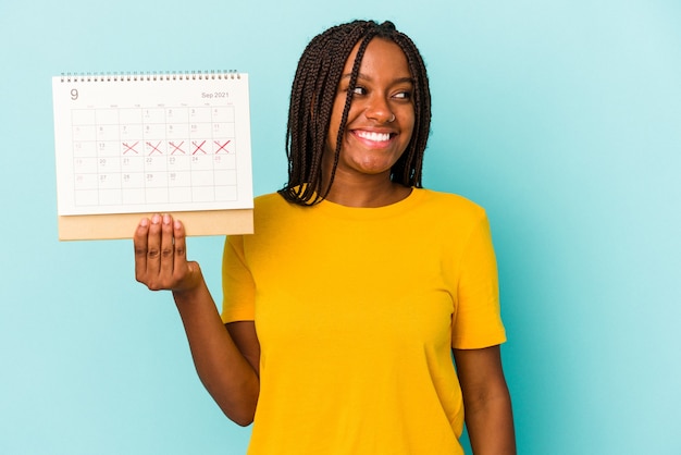 La giovane donna afroamericana che tiene un calendario isolato su fondo blu guarda da parte sorridente, allegra e piacevole.