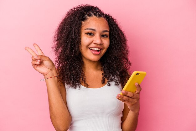 La giovane donna afroamericana che tiene un telefono cellulare ha isolato una parete rosa gioiosa e spensierata che mostra un simbolo di pace con le dita.