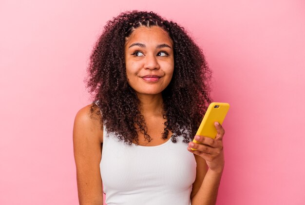 La giovane donna afroamericana che tiene un telefono cellulare ha isolato uno sfondo rosa che sogna di raggiungere obiettivi e scopi