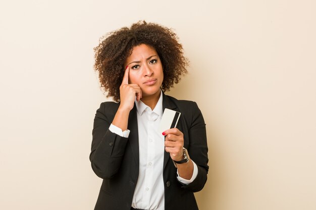 La giovane donna afroamericana che tiene una carta di credito che indica il suo tempio con il dito, pensando, si è concentrata su un compito.