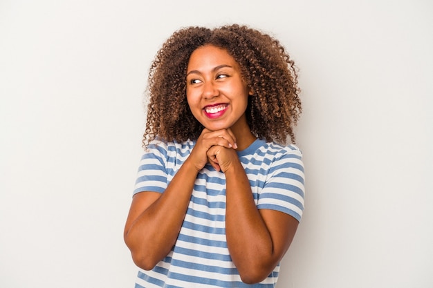 La giovane donna afroamericana con capelli ricci isolati su fondo bianco tiene le mani sotto il mento, sta guardando felicemente da parte.