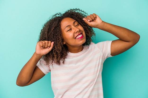 La giovane donna afroamericana con capelli ricci isolati su fondo blu che celebra un giorno speciale, salta e alza le braccia con energia.