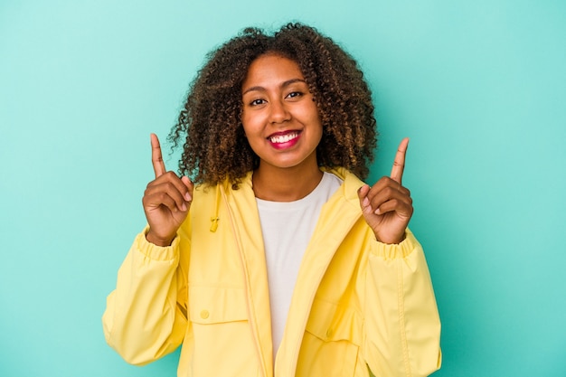 La giovane donna afroamericana con capelli ricci isolati su fondo blu indica con entrambe le dita anteriori in su mostrando uno spazio vuoto.