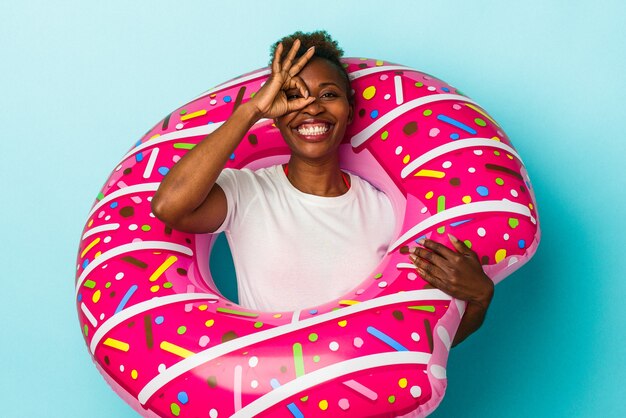 La giovane donna afroamericana con la ciambella gonfiabile isolata su fondo blu ha eccitato mantenendo il gesto giusto sull'occhio.