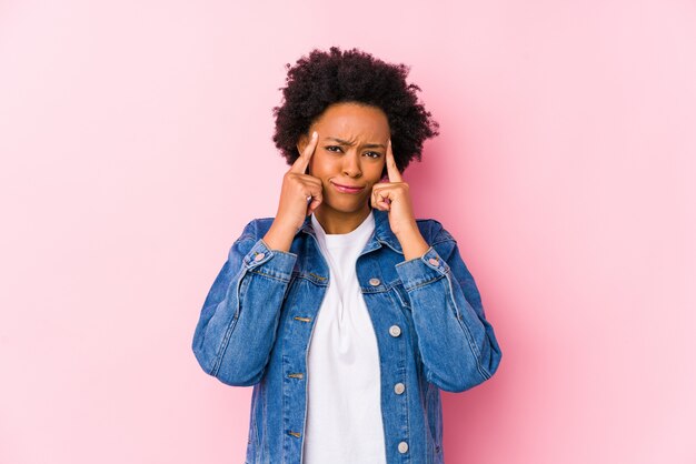 La giovane donna afroamericana contro un backgroound rosa isolata ha messo a fuoco su un compito, tenendo gli indici che indicano la testa.