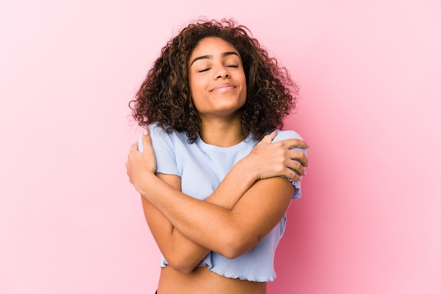 La giovane donna afroamericana contro un muro rosa abbraccia, sorridendo spensierata e felice.